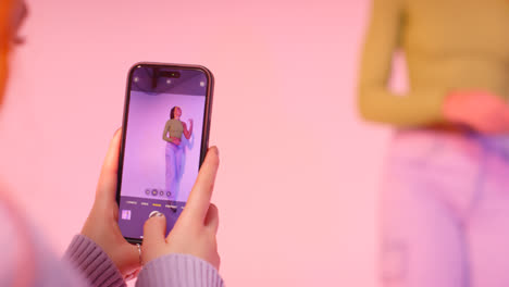 Studio-Shot-Of-Woman-Taking-Photo-Of-Friend-Dancing-On-Mobile-Phone-Against-Pink-Background-1